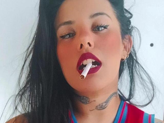 Yolisolange with black hair, red lipstick, and a cigarette in her mouth, wearing a red and blue top, has tattoos on her neck. She's looking slightly to the side.