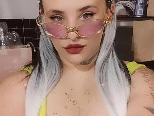Yolisolange with pink-tinted glasses, white-blue hair, red lipstick, tattoos, and a yellow top, showing her tits and freckles.