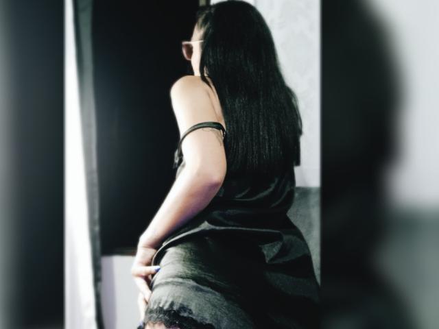 AnaisHilton with black hair, facing away, wearing a black top and dark striped skirt, showing her ass, standing in a shadowy, partially lit room.