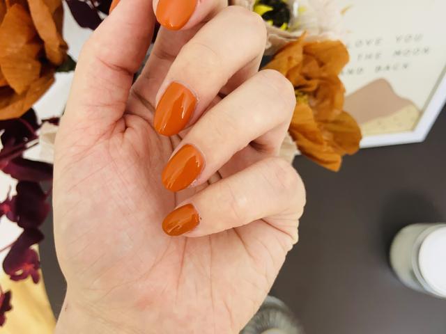 KarKutie's hand with shiny, orange-painted nails, showing off neat, oval-shaped tips. Her fingers are slightly curled, and the background has some blurred, yellow-orange flowers.