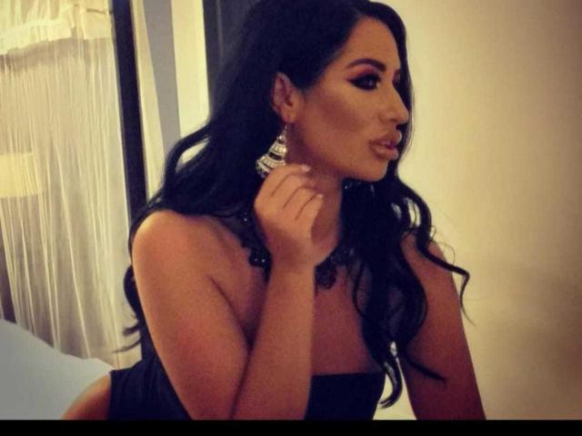 SexyAlyssa with long black hair, wearing a strapless black top, looking to the side, pouting lips, holding her neck, wearing large silver earrings, in a dimly lit room.