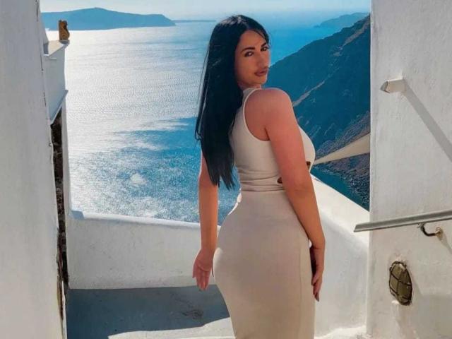 SexyAlyssa with long black hair, looking over her shoulder, wearing a tight beige dress, highlighting her big ass and curvy body, standing on a sunlit white balcony.
