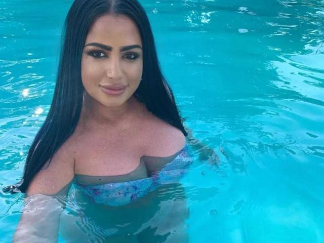 SexyAlyssa with long black hair, blue floral bikini top showing her big tits, and tan skin, standing in a clear blue pool, smiling at the camera.