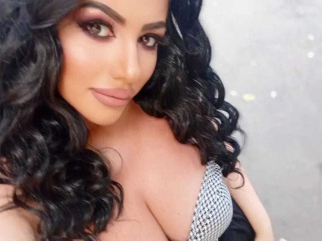 SexyAlyssa with curly black hair, full eyebrows, and pink lipstick, wearing a black-and-white houndstooth top, showing large, cleavage-filled tits. She's looking at the camera with a slight smile.