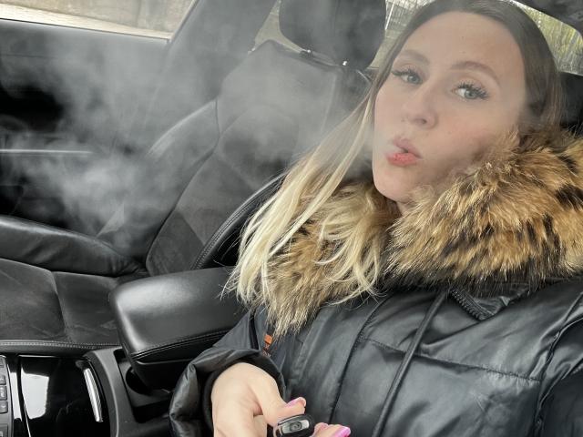 Saveta mit blonden Haaren, voller Lippen und blauen Augen, trägt eine schwarze, gepolsterte Jacke mit einer Fellkapuze und sitzt in einem verrauchten Auto, wobei sie ein pinkes Objekt hält..