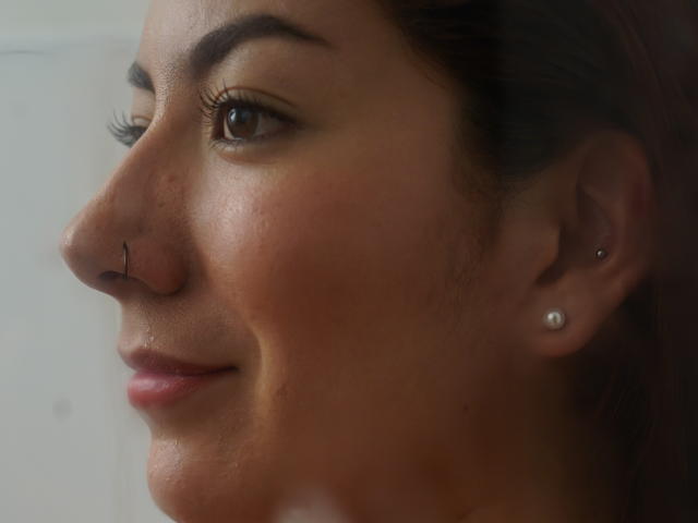 Het gezicht van AbbySton, met een neusring, bruine ogen en een lichte glimlach, met een parel oorbellen in haar linker oor. Haar huid is glad en ze kijkt naar de zijkant..