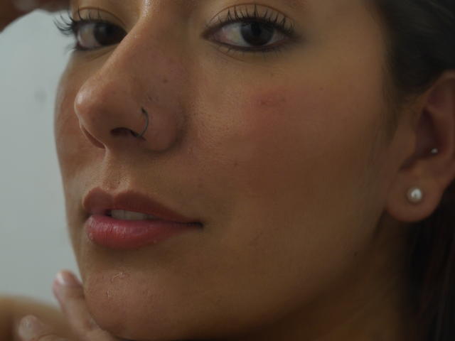 Het gezicht van AbbySton, met een neusring, lichtroze lippenstift, lichtrode wangen en een parel oorbellen, met haar hand in de buurt van haar kin. Haar ogen kijken naar de zijkant..