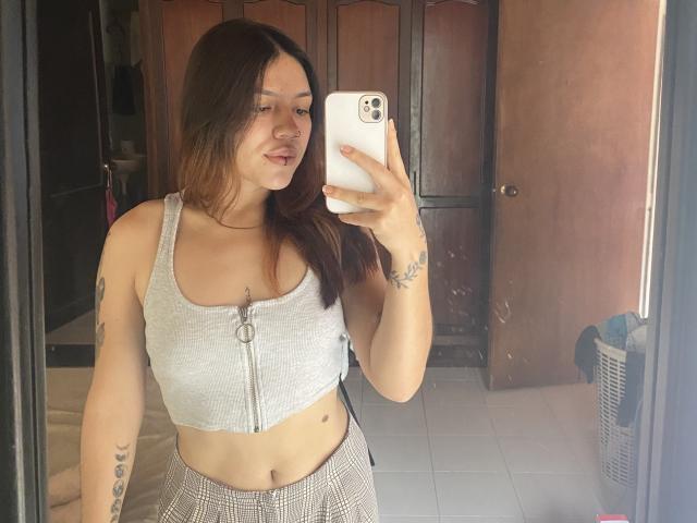SophiaMarx, com cabelo castanho, blusa cropped cinza com zíper, mostrando os seios, tatuagens nos braços, segurando um celular, saia xadrez, lábios curvados para baixo, selfie no espelho, portas de madeira ao fundo..
