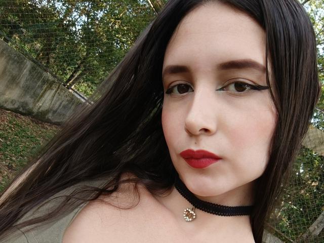 RacheFoxx has long dark hair, red lipstick, black choker with a gold flower, and green eyeliner. Her boobs are not visible. She's outdoors with trees in the background.