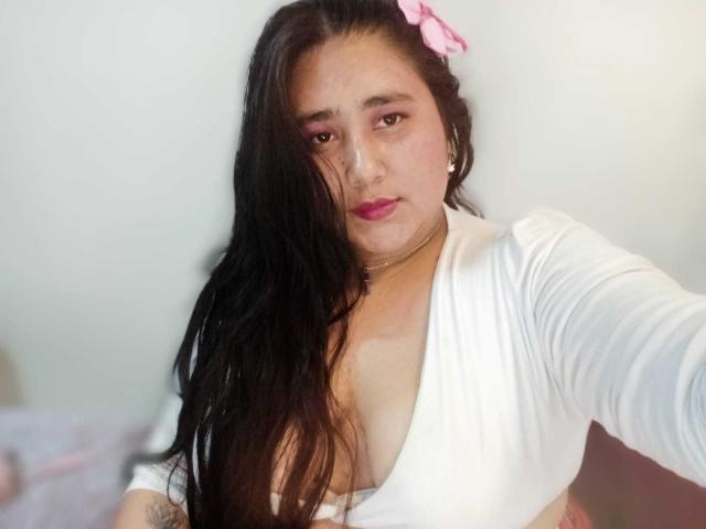 ZoeGiil with long black hair, pink flower, white deep V-neck top, showing her tits, pink eyeshadow, pink lipstick, small tattoo on right arm, taking a selfie.