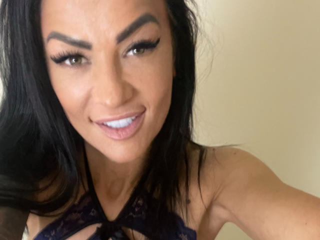 AndraD has dark, wavy hair, brown eyes, thick eyebrows, and is smiling with blue lipstick. She's wearing a black lace top, showing her cleavage.