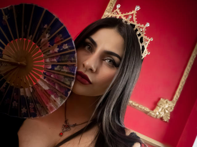 LombanaX with long black hair, red lipstick, black lace top, and denim shorts, sitting on a blue velvet chair, wearing a tiara, black stockings, and showing her tits.