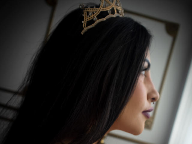 LombanaX with dark hair, wearing a gold crown, red lipstick, and a red dress, covering her tits with her hands, dark nail polish, red curtain, and jewelry.