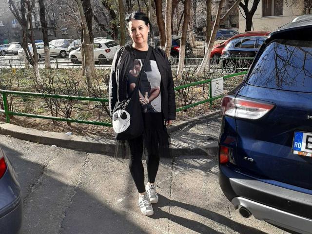 LolaGreate has short black hair, wears a black and gray print shirt, black cardigan, black pants, white sneakers, and a white crossbody bag, standing by a blue car.