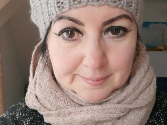 LolaGreate with a nose piercing, wearing a gray knit hat and beige scarf, has brown eyes with black eyeliner, a slight smile, and a light skin tone.
