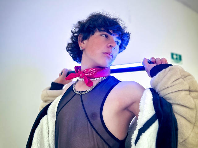 Luke Howlandy, con i capelli castano ricci, indossa una maglia nera trasparente che rivela un piccolo pene flaccido, una bandana rossa, una collana a catena e una giacca beige, e tiene una barra blu sulla spalla..