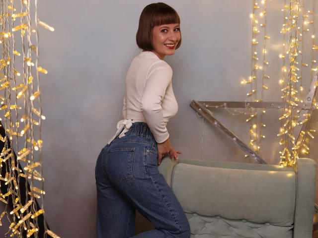 LizzyWest with a cute bob, smiling, wearing a white long-sleeve top and high-waisted blue jeans, leaning on a green couch, showcasing her round ass. String lights in background.