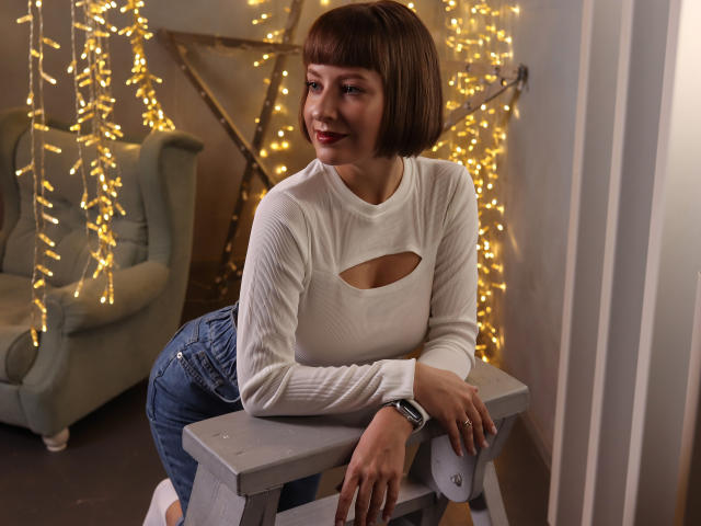 LizzyWest with short brown bob, white long-sleeve top with cutout, blue jeans, leaning on gray table, smiling, wearing black watch, surrounded by yellow string lights.