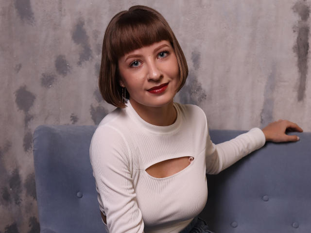 LizzyWest with short brown bob, white cutout top, blue eyes, red lipstick, smiling, sitting on blue couch, right arm on seat, background gray and textured.