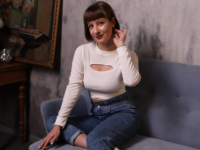 LizzyWest with short brown hair, white long-sleeve top with cutout, showing small tits, blue jeans, sitting on blue sofa, smiling, right hand touching ear.