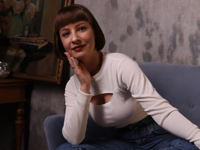 LizzyWest with a cute bob, wearing a white long-sleeve top with a cutout, sitting on a blue couch, hand on cheek, smiling, red lipstick, denim jeans, ring.
