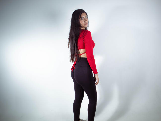 KarinaVughet with long black hair, red crop top with cutouts, and black leggings, standing with her back to the camera, looking over her shoulder, showing her ass.