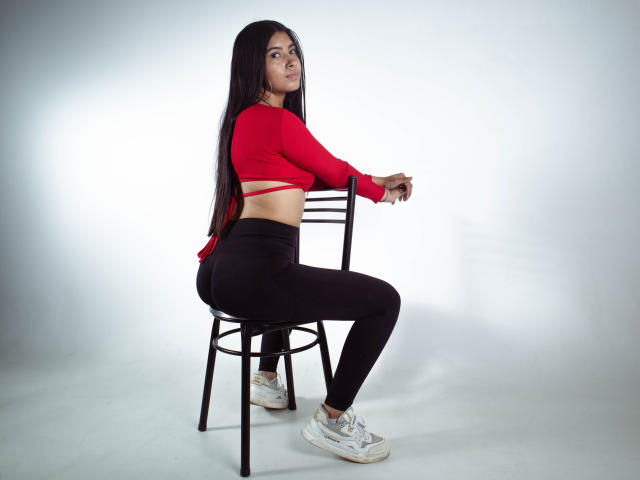 KarinaVughet, com cabelos longos e pretos, uma blusa cropped vermelha, calças legging pretas e tênis brancos, sentada de lado em uma cadeira, mostrando seu traseiro e uma faixa vermelha sob a blusa..