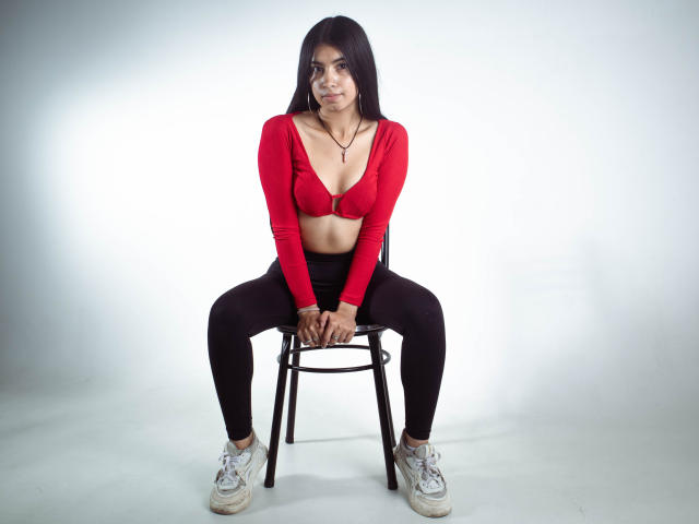 KarinaVughet, con cabello largo y negro, un top rojo que deja ver sus senos, pantalones negros y zapatillas blancas, sentada en una silla con las piernas separadas y las manos sobre las rodillas..
