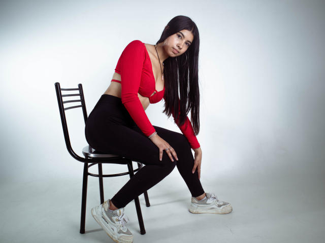 Karina, avec de longs cheveux noirs, un haut court rouge, un pantalon noir et des baskets blanches, assise sur une chaise noire, penchée en avant, les mains sur les genoux, regardant la caméra..