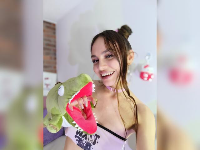 LiliiMoore with brown hair in a bun, smiling, holding a green shark stuffed toy with a red open mouth. She wears a pink choker and a white top.