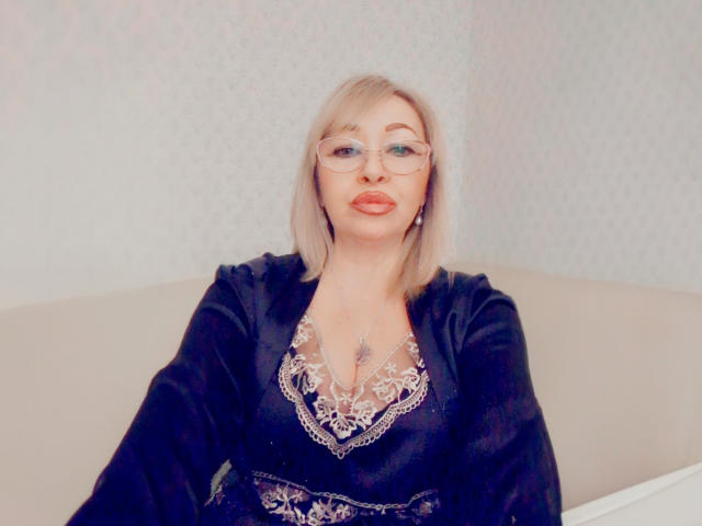 Lori, con i capelli biondi, gli occhiali e le labbra rosse, indossa un abito di velluto nero sopra una camicetta con pizzo, seduta su un divano beige. Il seno è visibile attraverso il pizzo..
