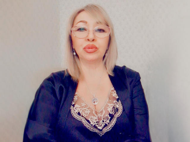 LoriExcite, con cabello rubio, gafas y lápiz labial rojo, vestida con un vestido negro con un estampado de encaje, que muestra su pecho, y un collar de plata, tiene labios rojos y voluminosos..