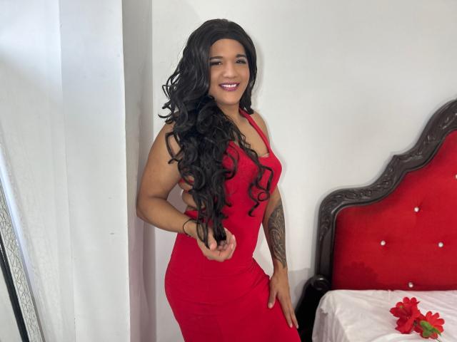 Un uomo con capelli neri ricci e lunghi, vestito rosso, sorridente, in piedi in una camera da letto, che mostra un tatuaggio sul braccio destro, un letto rosso con un cuscino bianco, un fiore rosso sul letto..