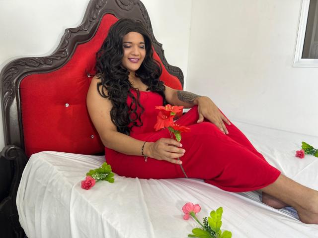 Una donna con lunghi capelli neri ricci, vestita con un abito rosso, seduta su un letto con una testiera rossa imbottita, che tiene un fiore rosso, con rose rosa sparse intorno..