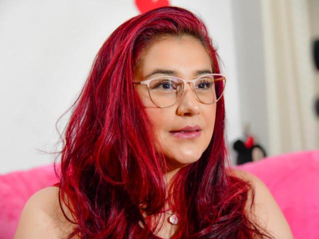 JadeSweetSweet has red hair, wears glasses, and looks at the camera with a slight smile. She has light skin, pink lips, and a necklace. Boobs not visible.