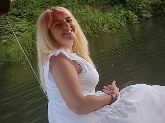 LisaXSienna with blonde hair, smiling, wearing a white dress, sitting by a green river, holding her hands, showing her tits, with a bracelet on her wrist.