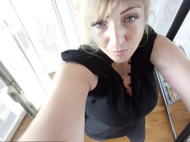 LisaXSienna with blonde hair and blue eyes, wearing a black sleeveless top, showing her tits in a selfie, standing near a window with wooden floor.
