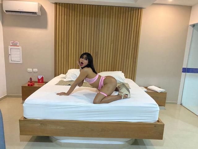 NairoBits, with pink lingerie and high heels, is on all fours on a bed, showing her ass and tits, with a surprised expression, in a modern bedroom.