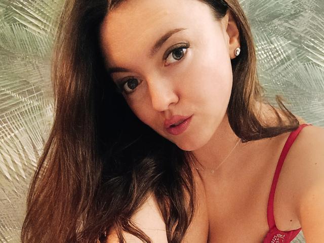Vanilia69 with long brown hair, green eyes, and red bra, showing off her tits and slightly parted lips, wearing a delicate necklace, against a textured silver background.