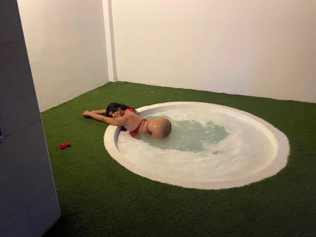 A AnnaGrey está em uma pequena banheira de hidromassagem circular, deitada de bruços com o traseiro para cima, vestindo um biquíni vermelho, em um tapete verde. Sua bunda molhada é bem visível..