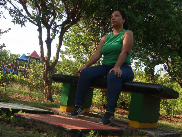 ElChicletv, with big tits, sits on a black green-yellow bench, wearing a green sleeveless top, blue jeans, and black sneakers, in a sunlit, wooded park.