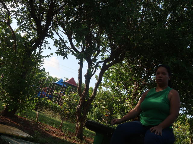 ElChicletv, a chubby woman with dark hair in a green sleeveless top, sits on a green metal bench, looking to the side, in a sunny, leafy park.