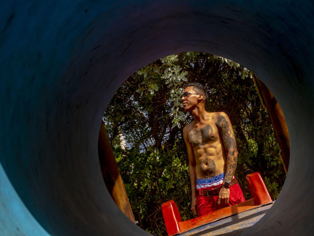 Shirtless ElChicletv with tattoos, wearing red shorts, stands in a circular blue tunnel, looking up with sunglasses on, sunlight highlighting his toned abs and dark skin.