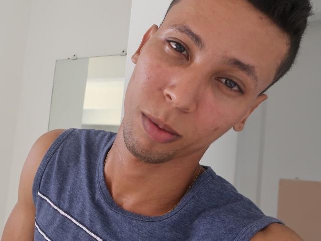 ElChicletv has a cute face with light brown skin, short black hair, and blue eyes. He's wearing a sleeveless blue shirt, looking slightly off-camera with a subtle smile.