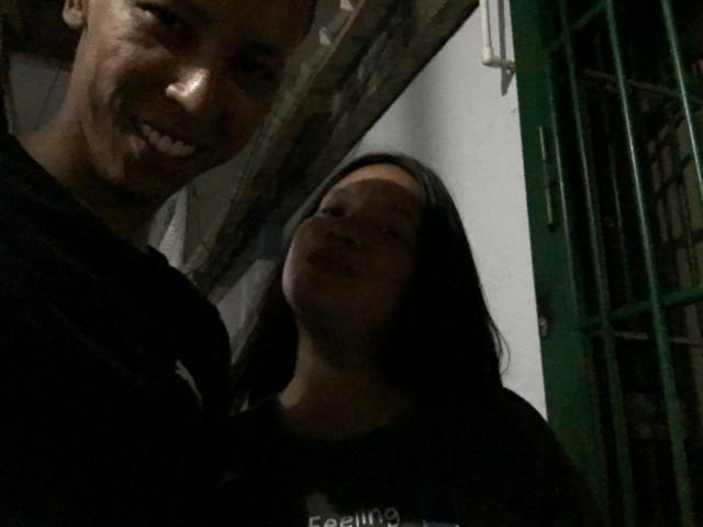 ElChicletv with dark hair and closed eyes, smiling, stands beside a taller, darker-skinned ElChicletv with a broad grin, both wearing black shirts in dim lighting.