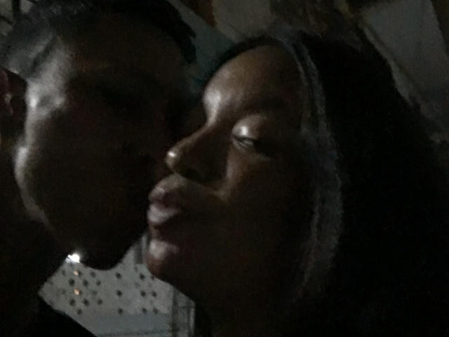 ElChicletv's dark face is next to a man's face in dim light, their noses almost touching, both with slightly open mouths, in a low-light, grainy, intimate shot.