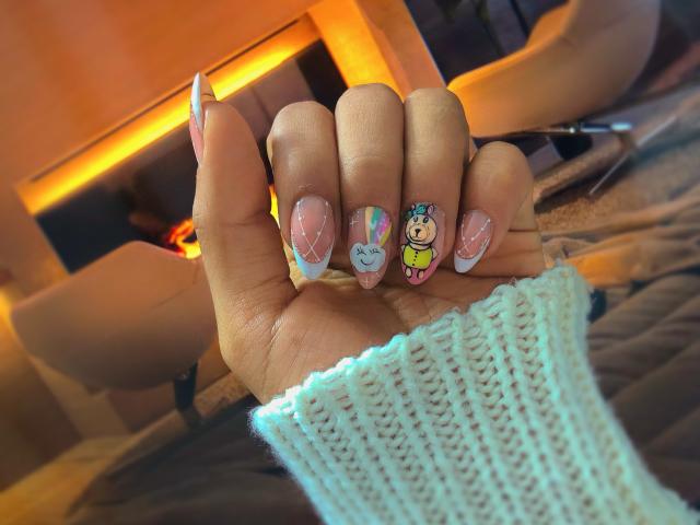 Lunas Hand mit farbenfroher Nagelkunst: Pastellrosa mit Diamantmustern, eine blaue, lächelnde Katze und ein gelbes Cartoon-Figuren. Sie trägt einen weißen, gestrickten Ärmel..