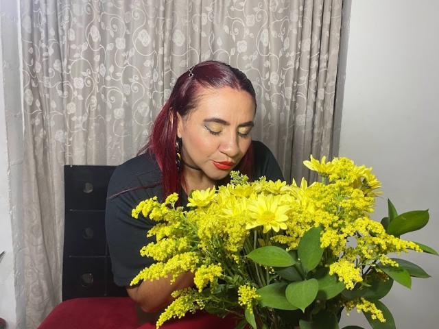 NahelaBigMelonsX with red lipstick and dark hair, holding a large bouquet of yellow flowers, eyes closed, sitting on a red chair, wearing a black shirt.