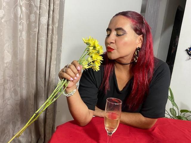 NahelaBigMelonsX with red hair, black shirt, holding yellow flowers, nose close, red lipstick, gold earrings, bracelet, glass of white wine on red tablecloth, gray curtains.