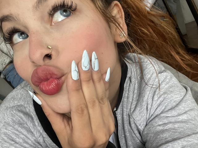 Shantal, com olhos azuis, batom vermelho e unhas brancas em formato de garras, fazendo beicinho, usando um piercing no nariz e um moletom cinza com zíper, olhando para cima..